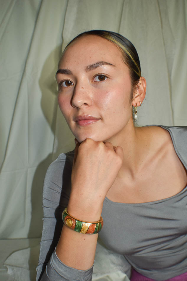 Woman wearing a gray top with a green and gold bracelet, sitting against a plain background.