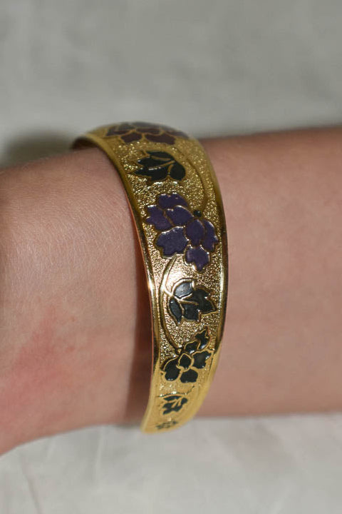 Gold bracelet with purple and green design on a wrist against a neutral background