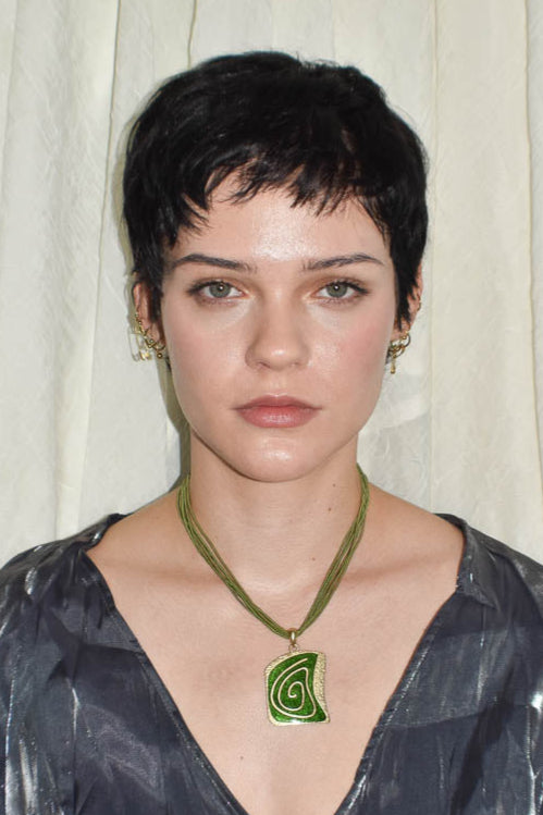 Woman wearing a dark patterned top and green necklace with a leaf design against a white curtain background